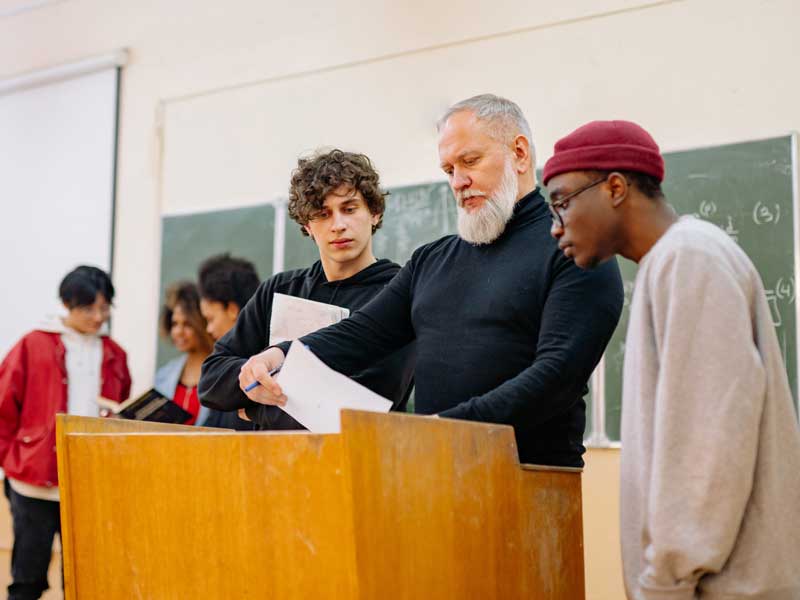 students reviewing papers with a professor at a classroom podium